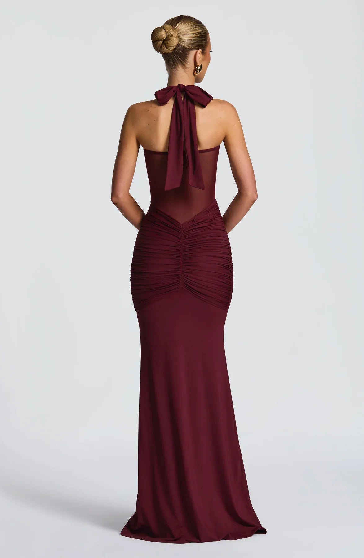 Back view of a burgundy halter neck maxi dress with a tied bow and elegant ruched detailing on the rear.