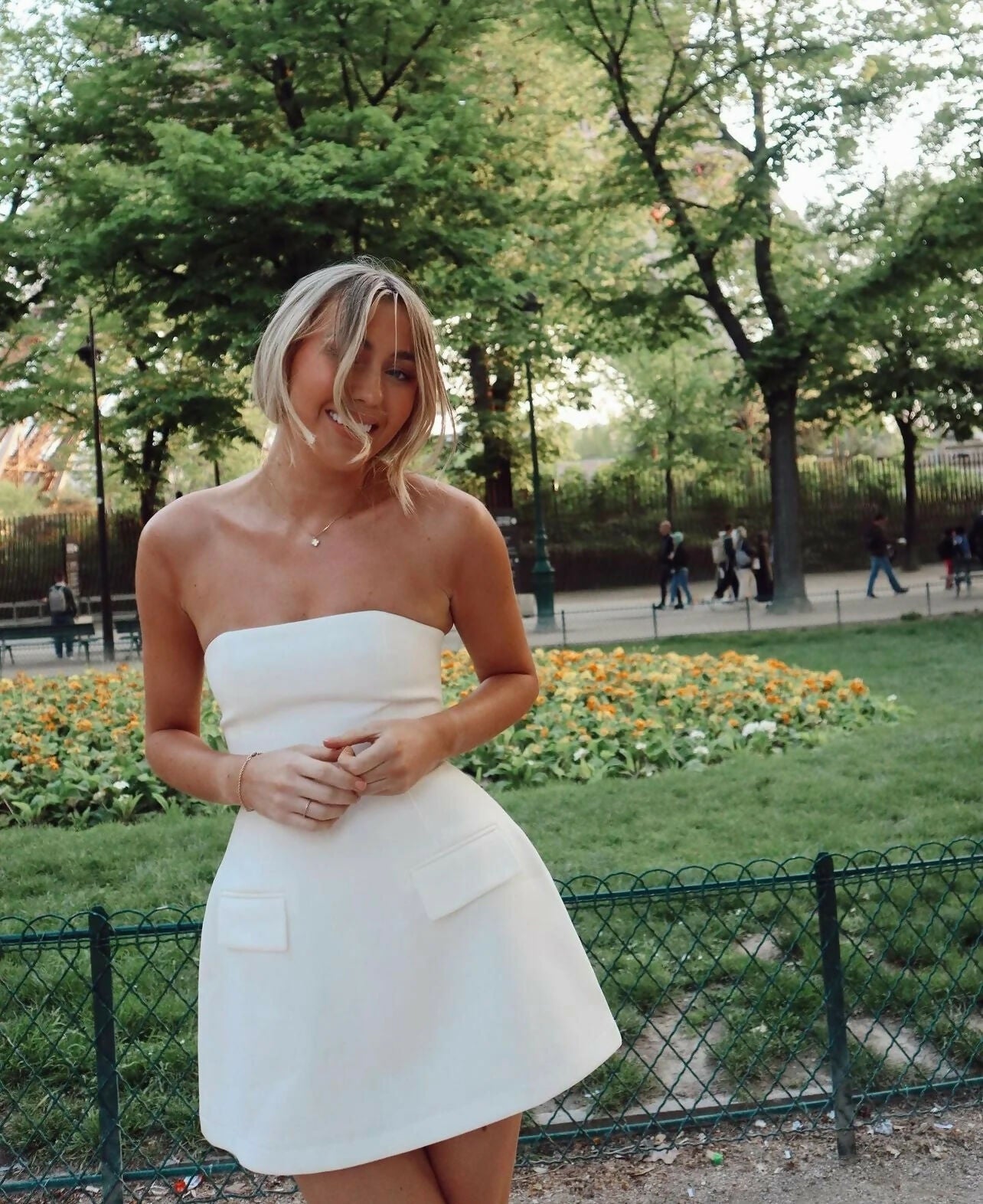 Smiling woman in a white strapless mini dress with flap pockets and cinched waist, standing in a park setting.