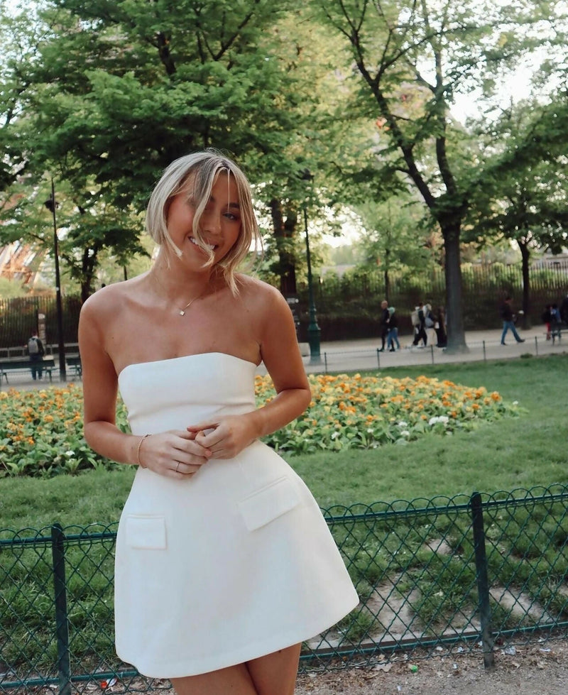 Smiling woman in a white strapless mini dress with flap pockets and cinched waist, standing in a park setting.
