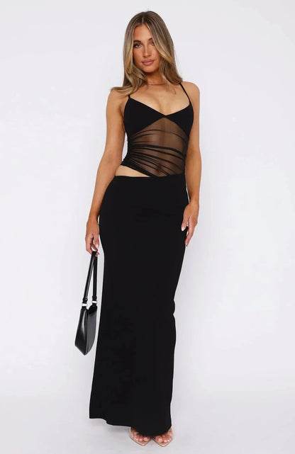 Front full-body shot of a woman in a black asymmetrical cutout ruched mesh maxi dress with a halter neckline.