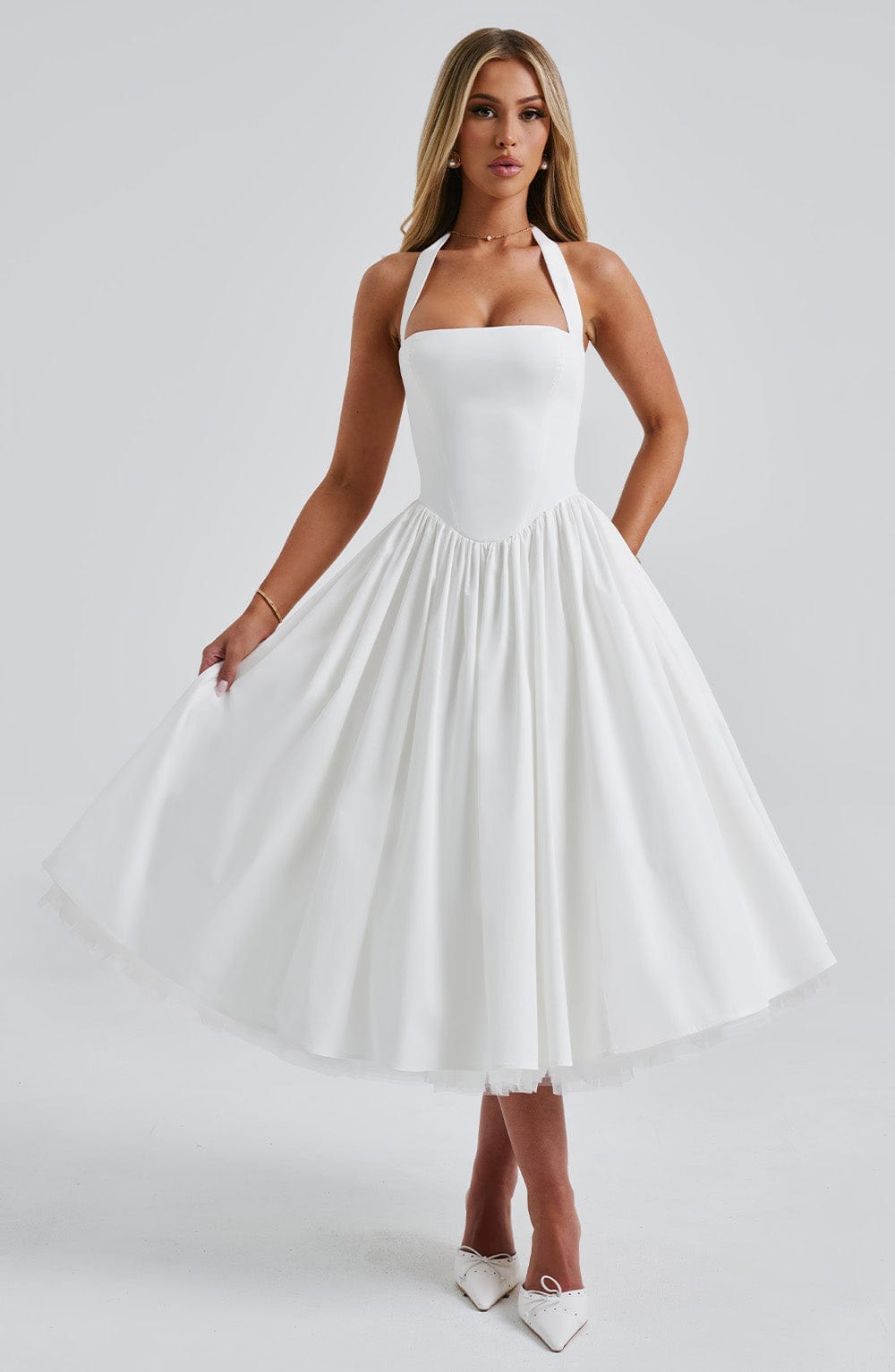 White backless halter neck midi dress with a full skirt and visible tulle underskirt, shown with hands gently lifting the hem.