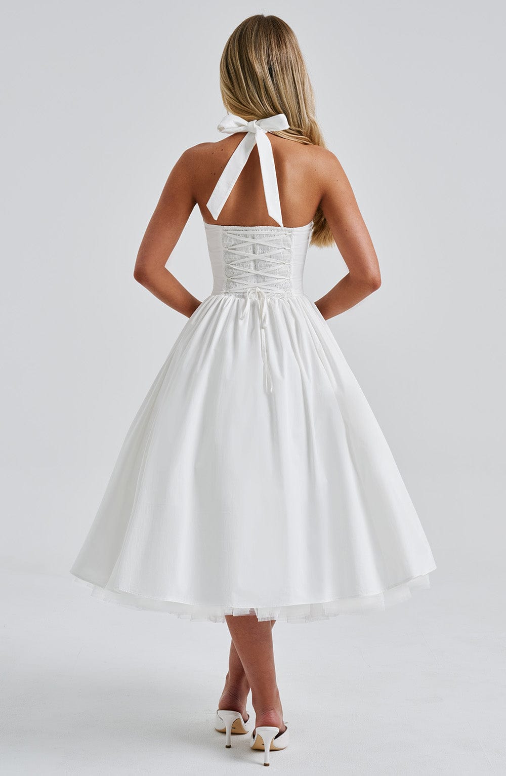 Back view of a white backless halter neck midi dress with a lace-up corset detail and tie closure at the neck.