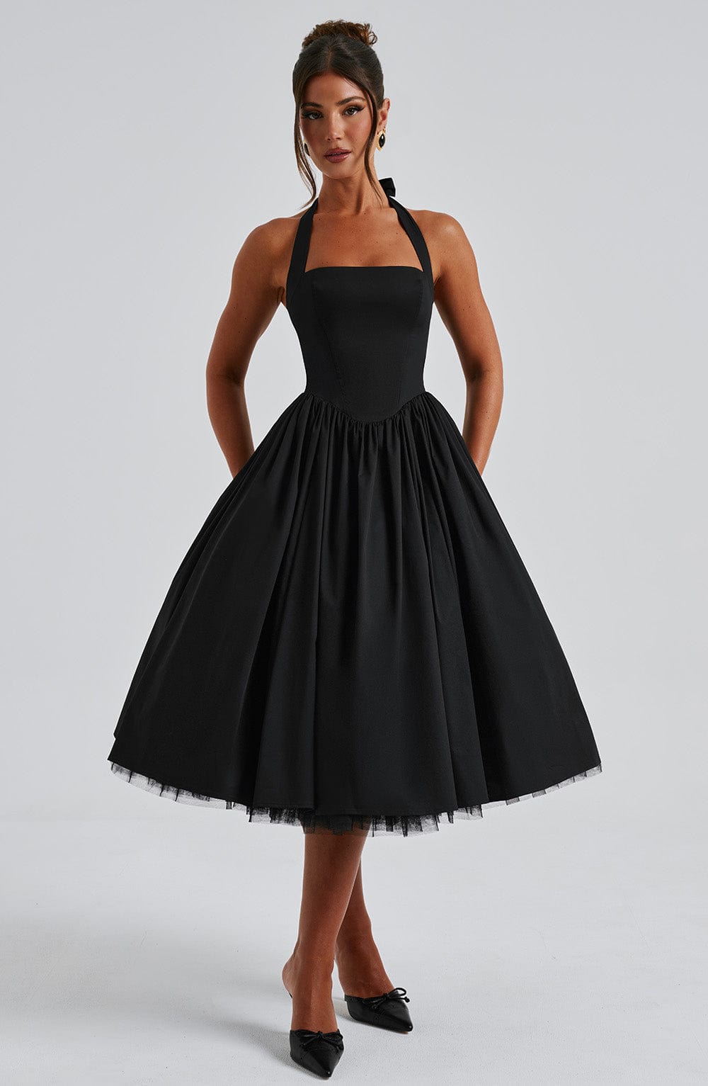 Black backless halter neck midi dress with a voluminous skirt and structured bodice, shown with hands in pockets.