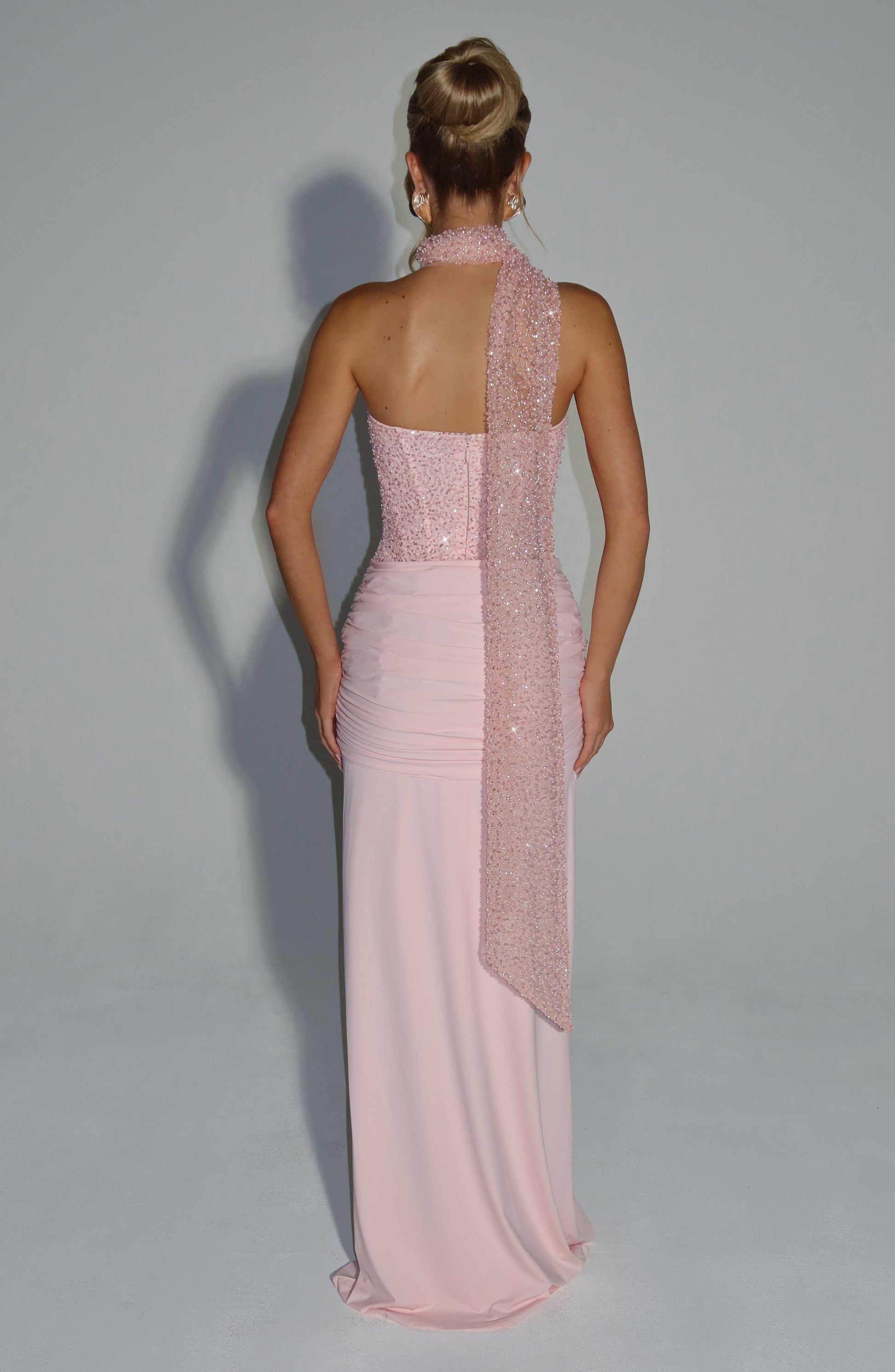 Back view of a blush sequin corset strapless maxi dress featuring a ruched skirt and a long sequin scarf draped over one shoulder.