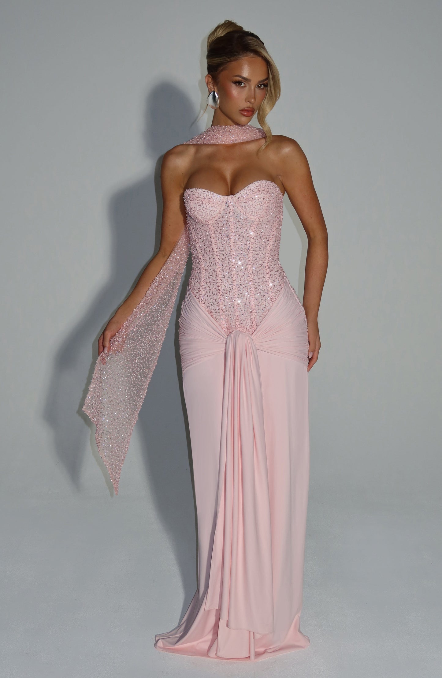 Front view of a blush sequin corset strapless maxi dress with a draped skirt and matching sequin scarf detail, perfect for wedding guests.