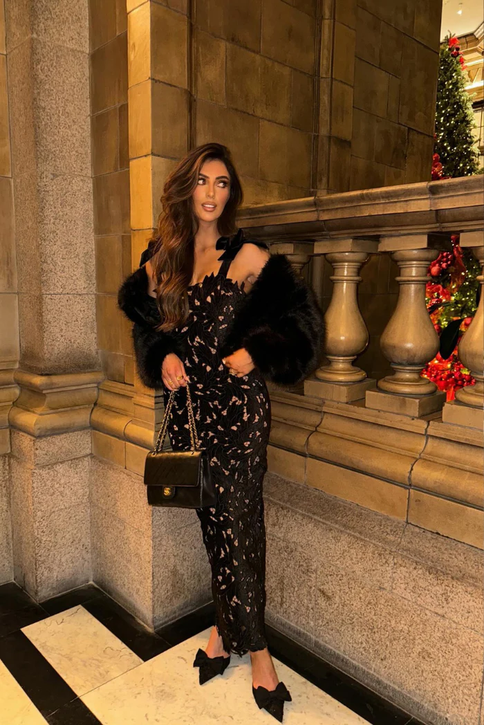 Woman standing indoors in a black bow shoulder lace bodycon maxi dress with a faux fur jacket and a black quilted handbag.