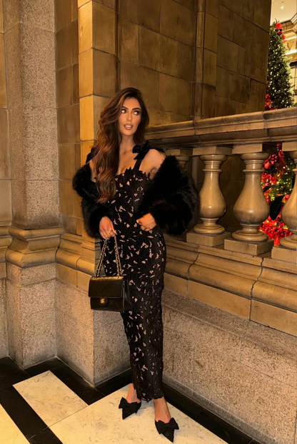Woman standing indoors in a black bow shoulder lace bodycon maxi dress with a faux fur jacket and a black quilted handbag.