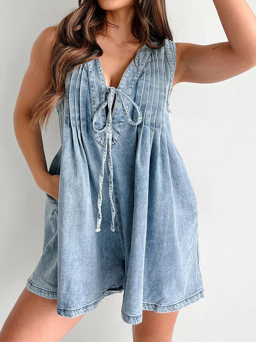 Light wash denim romper for women featuring a V-neckline with a tie-front detail and pleated bodice, showcasing a relaxed fit with pockets.