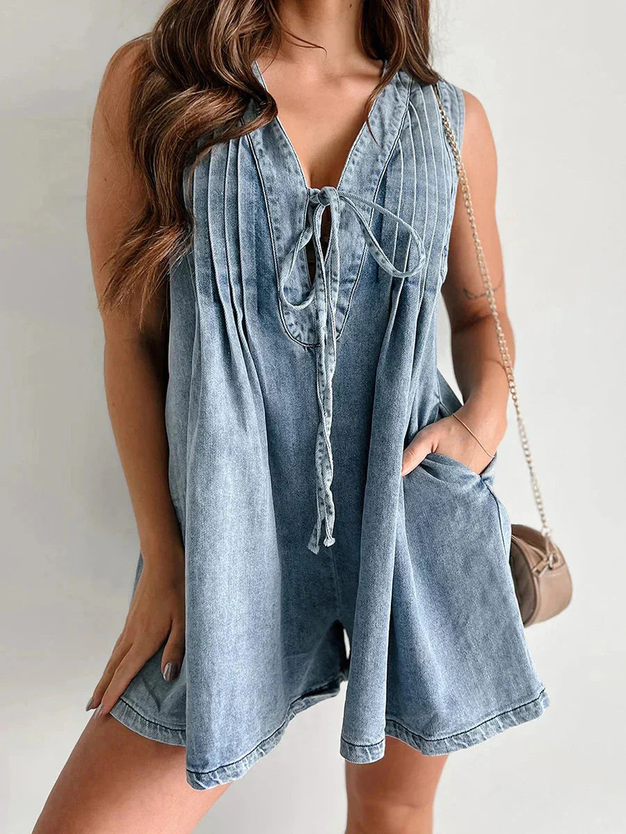 Women's light blue denim romper with a tie-front V-neck and pleated front, styled with a small cross-body bag and hands in pockets.