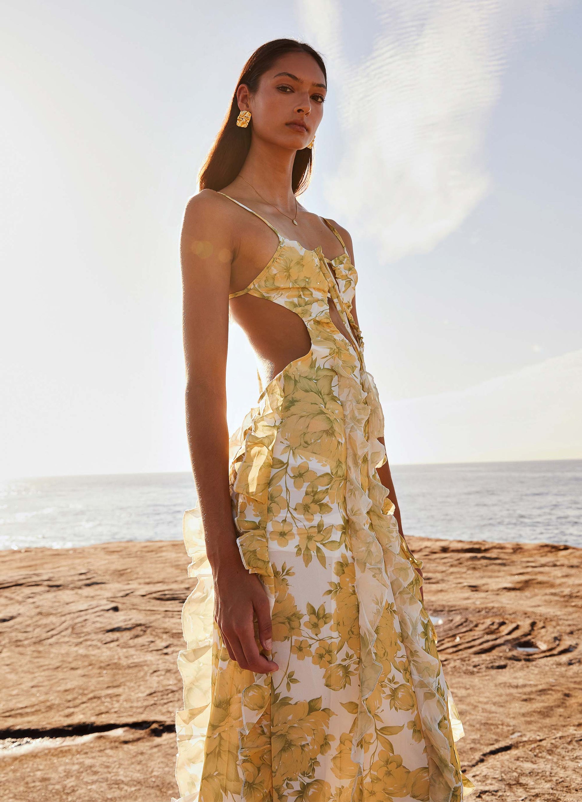 Women's floral cut out ruffle tie front maxi dress in yellow and white, standing on rocky beach, vacation dresses style.