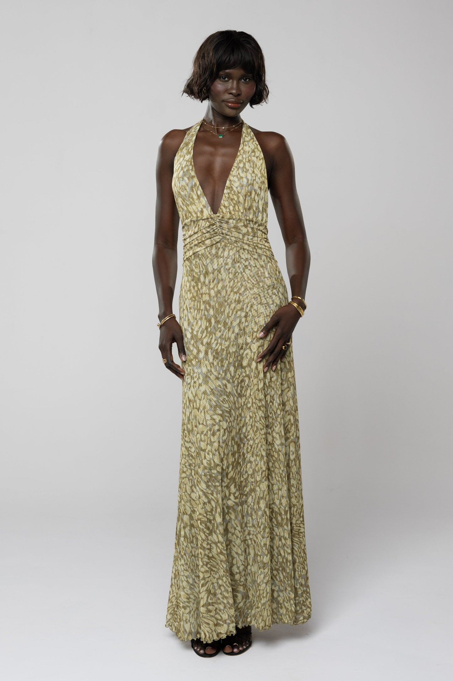 Women's halter maxi dress in a subtle green and yellow abstract floral print, with a deep v-neck and ruched waist, suitable for a special occasion.