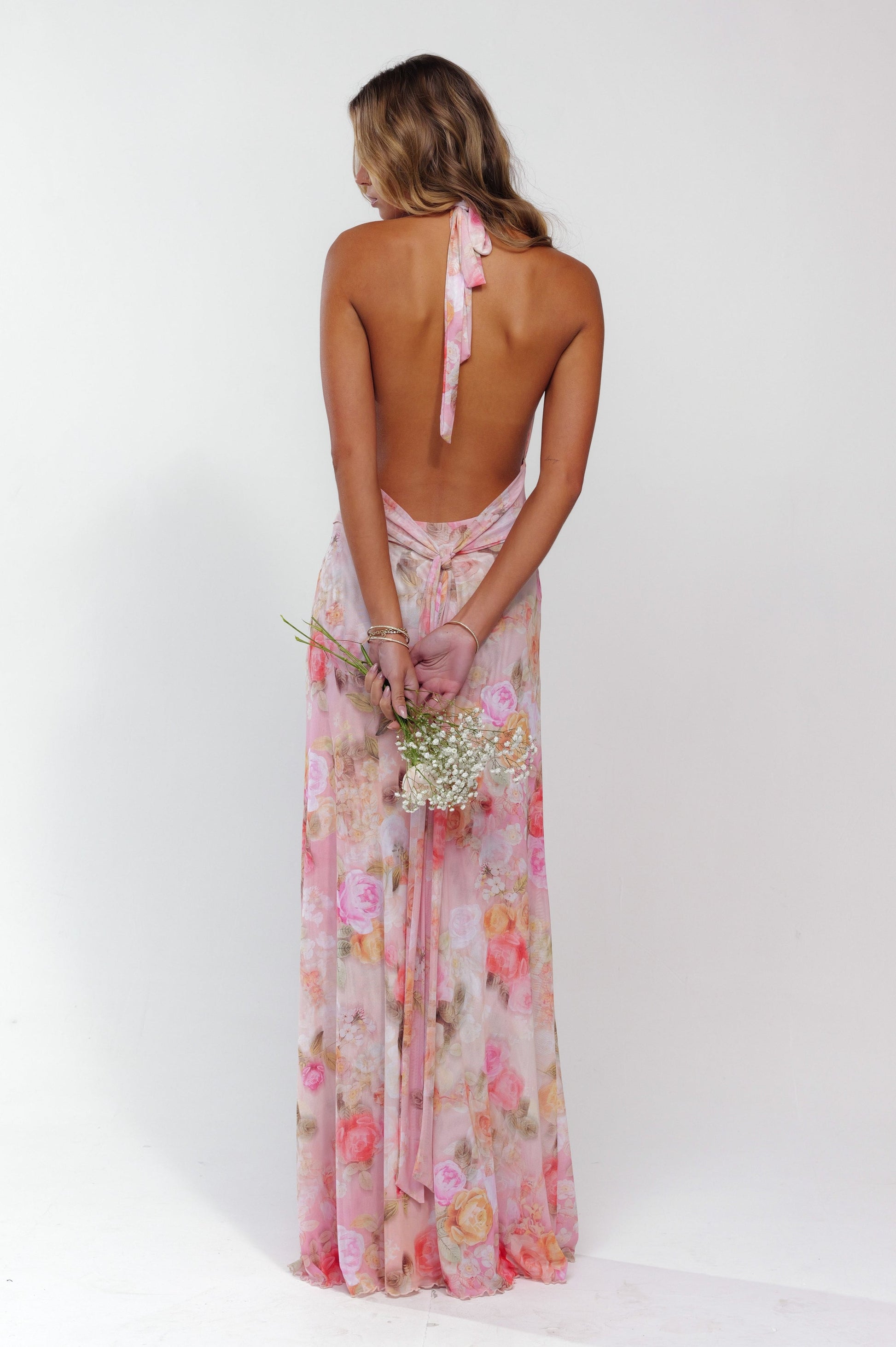 Rear view of a women's pink floral halter maxi dress, highlighting the open back and long skirt, holding flowers.