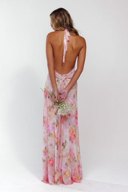 Rear view of a women's pink floral halter maxi dress, highlighting the open back and long skirt, holding flowers.