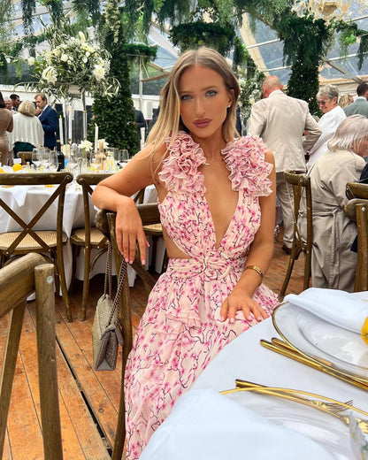 Women's summer floral V-neck maxi dress in light pink with white floral print, ruffled shoulders and waist cutouts, sitting at a table.