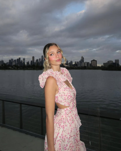 Side profile of a woman wearing a white and pink floral maxi dress, featuring a ruffled V-neckline and side cutouts, against a city skyline.