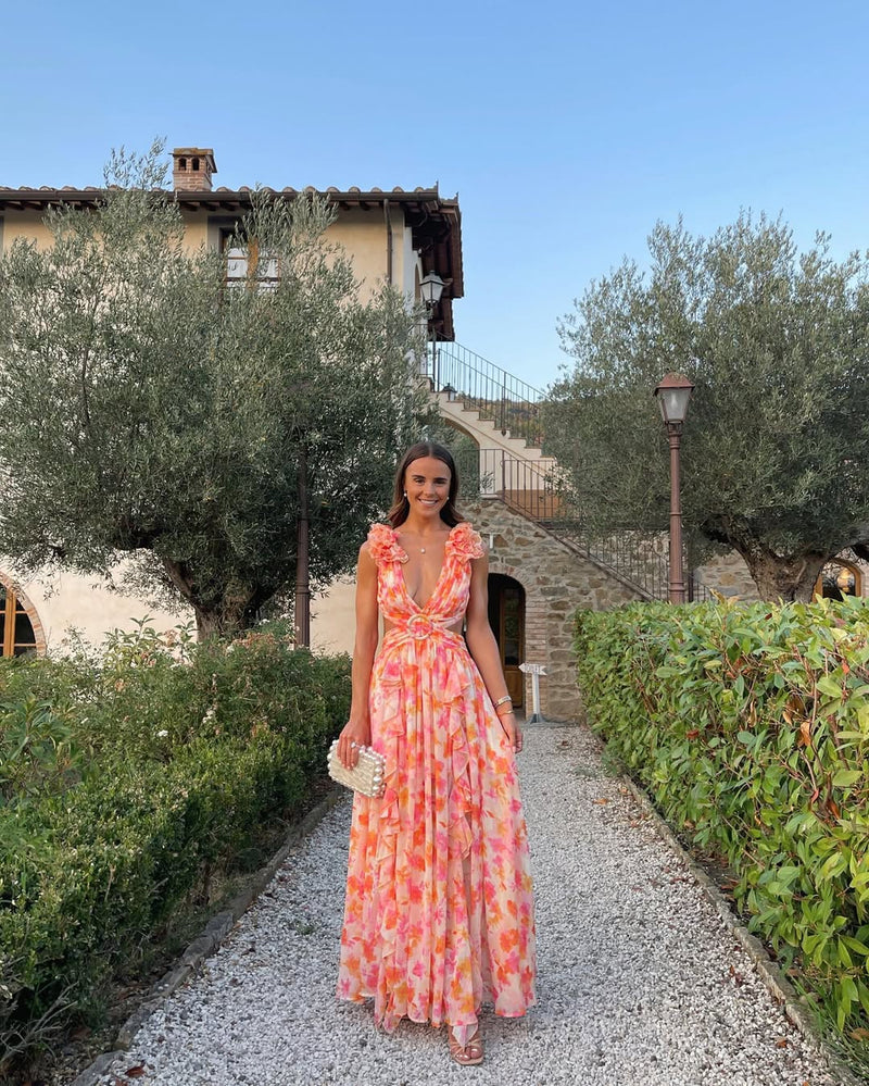 Women's summer floral V-neck maxi dress in vibrant orange and pink, with ruffled shoulder details, standing on a gravel path.