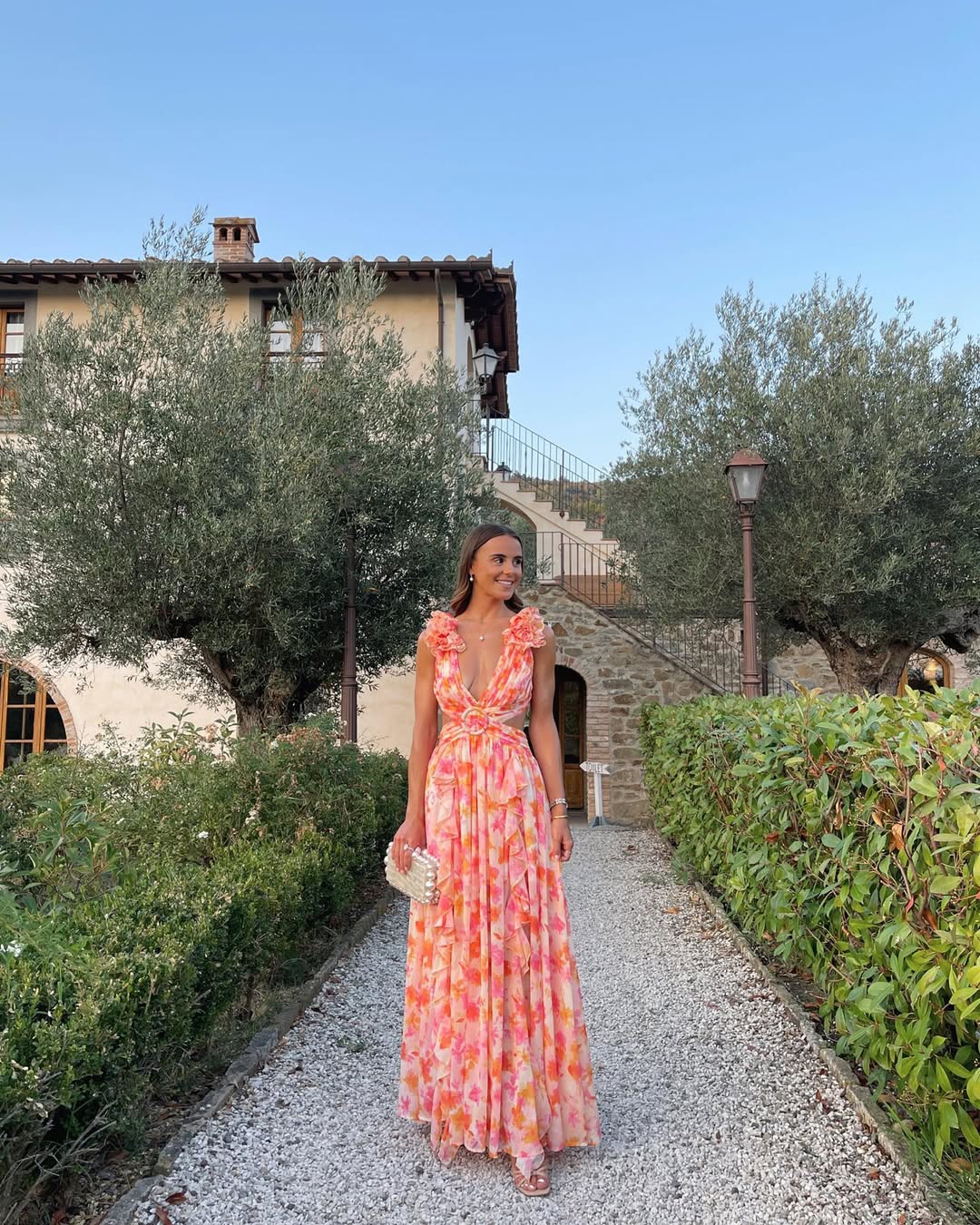 Women's summer floral V-neck maxi dress in orange and pink, with ruffled shoulder details, looking to the side on a gravel path.
