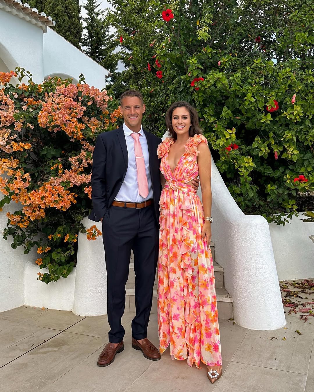 Couple posing, woman wearing a summer floral V-neck maxi dress in orange and pink, with ruffled shoulders and waist cutouts.
