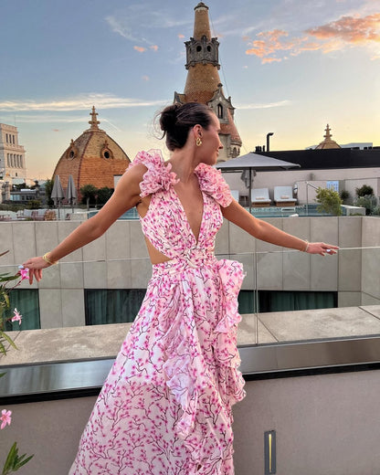 Women's summer floral V-neck maxi dress in light pink with white floral print, ruffled shoulders and waist cutouts, arms outstretched on a rooftop.
