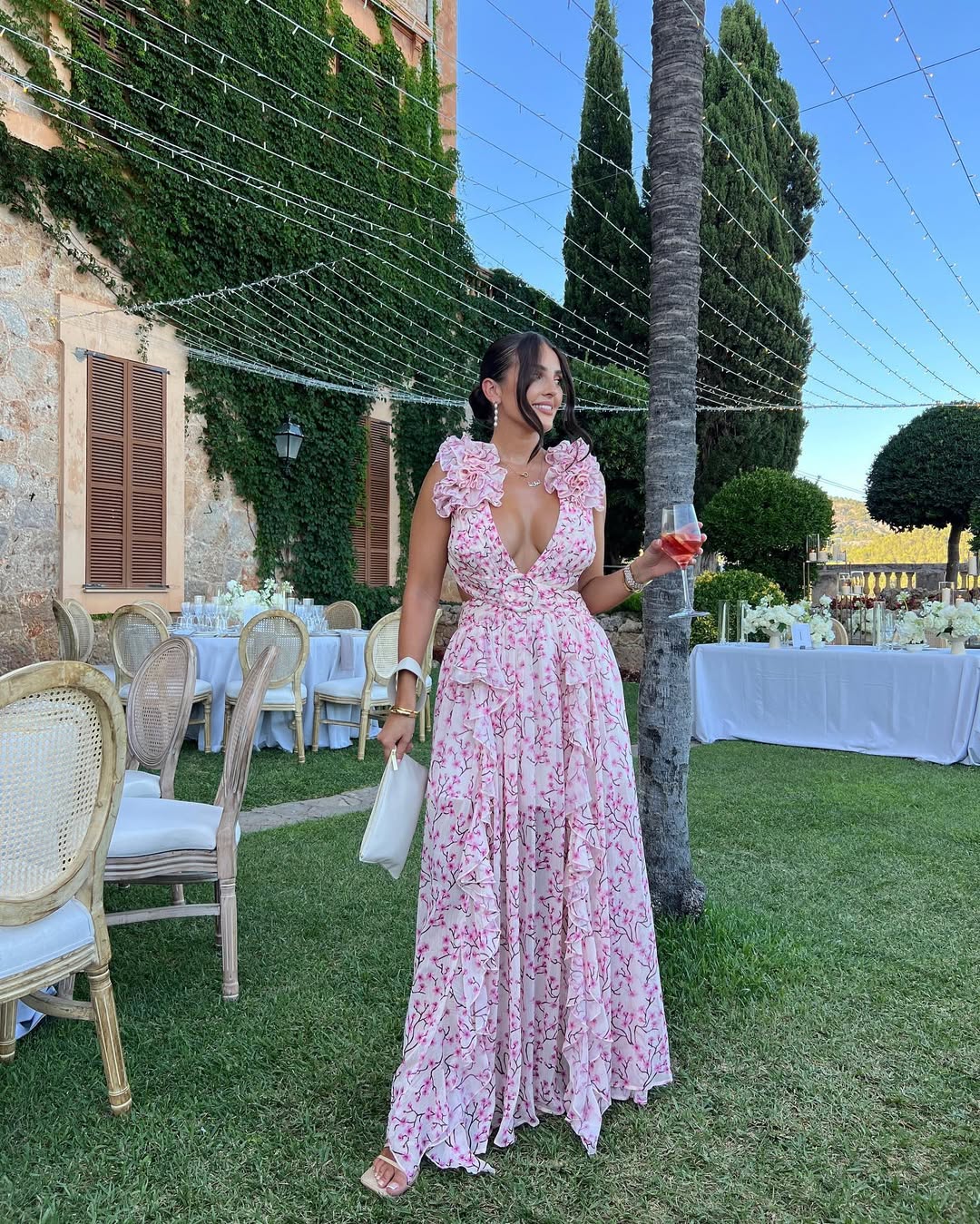 Women's summer floral V-neck maxi dress in light pink with white floral print, ruffled shoulders, holding a drink at an outdoor event.