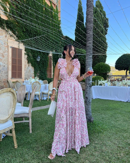 Women's summer floral V-neck maxi dress in light pink with white floral print, ruffled shoulders, holding a drink at an outdoor event.