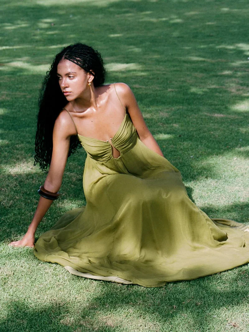 Women's olive green gathered bust keyhole maxi dress, with a flowing skirt, worn by a model sitting on green grass.