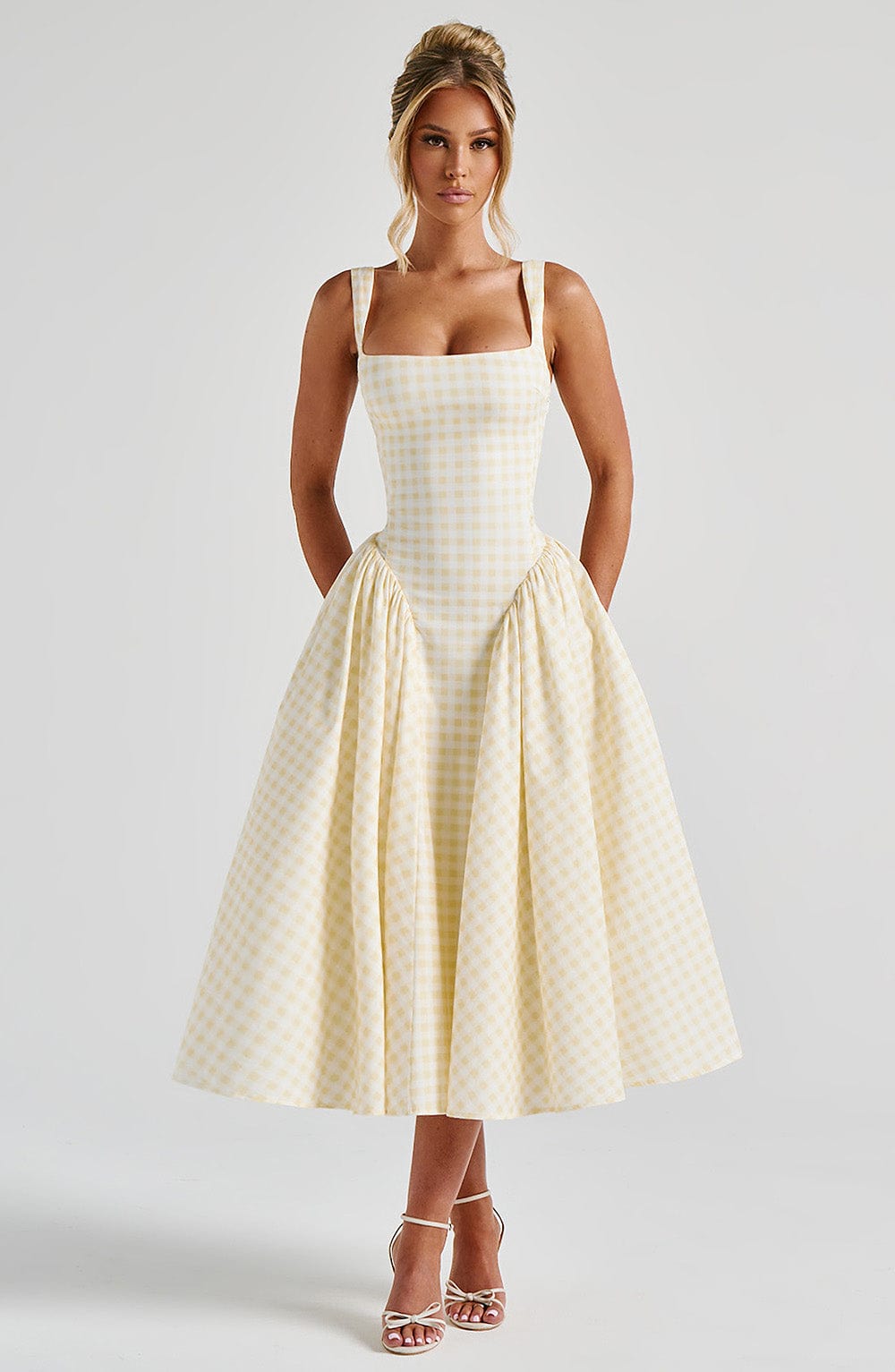 Woman wearing a light yellow gingham square neck midi dress with a fitted bodice and a full, pleated skirt, paired with white heels.