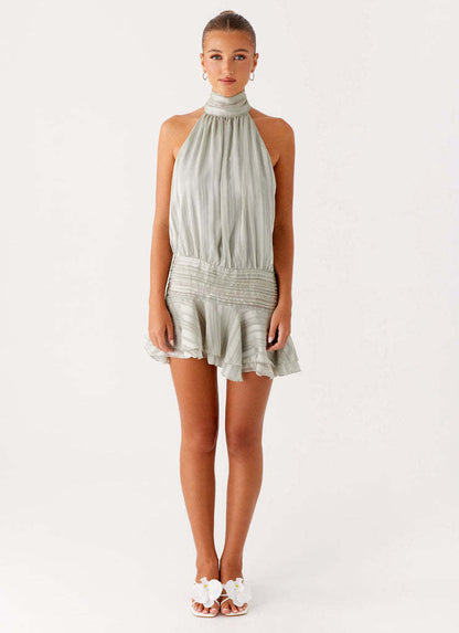 Full body view of a light sage green halter neck mini dress with a ruched waist, paired with white floral slides.