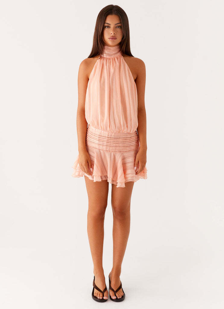 Full-length view of a women's peach halter neck mini dress with ruched waist and ruffled skirt, worn with black slides.