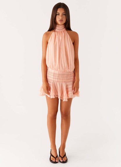 Full-length view of a women's peach halter neck mini dress with ruched waist and ruffled skirt, worn with black slides.