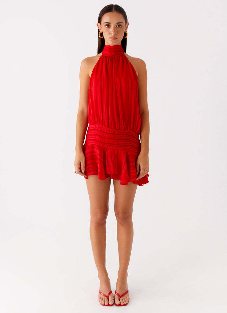 Full-length front view of a women's red halter neck mini dress with ruched waist and ruffled hem, worn with red slides.