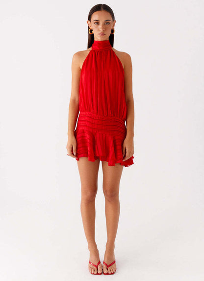 Full-length front view of a women's red halter neck mini dress with ruched waist and ruffled hem, worn with red slides.