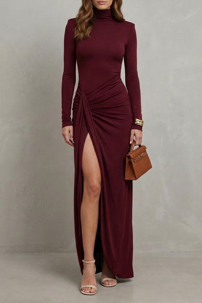 Burgundy long sleeve maxi dress with a high neck and draped slit, paired with a small handbag.