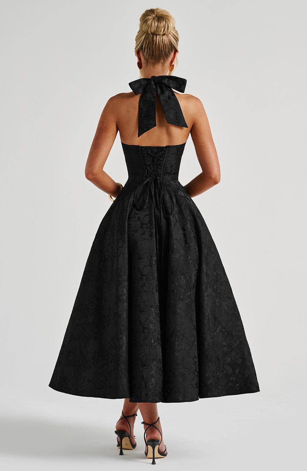 Rear view of a black jacquard halter neck midi dress, featuring a large bow and corset back detail.