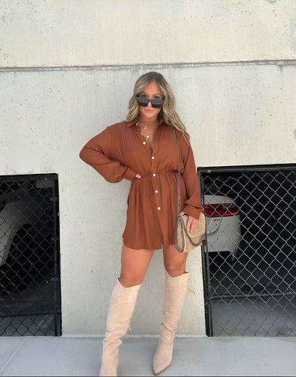Rust brown long sleeve button front mini romper with cinched waist, worn with tall beige boots and a crossbody bag.