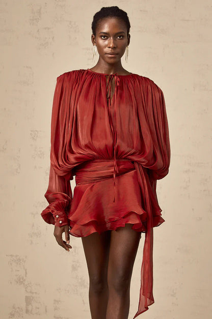 Full length shot of a woman in a vibrant rust-colored long sleeve tie neck mini dress, showing the draped fabric and pearl button cuffs.