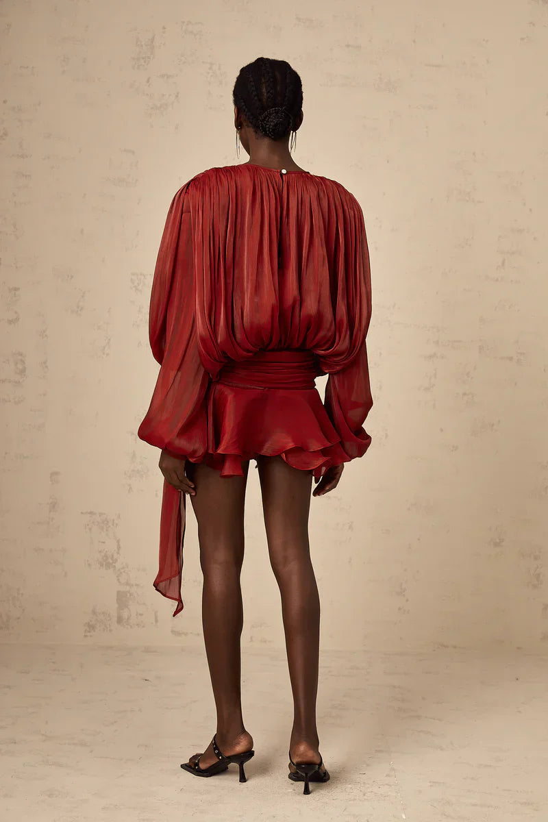 Back view of a women's long sleeve tie neck mini dress in a rich rust red color, featuring voluminous sleeves and a cinched waist with a long flowing belt.