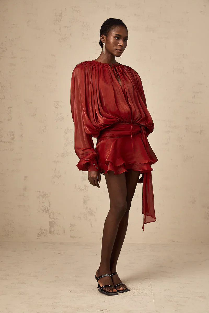 Front view of a women's long sleeve tie neck mini dress in rust red, showcasing the relaxed fit bodice, ruffled hem, and a self-tie belt detail.