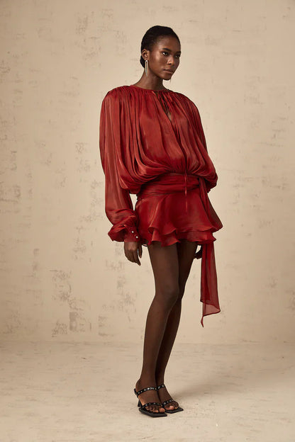 Front view of a women's long sleeve tie neck mini dress in rust red, showcasing the relaxed fit bodice, ruffled hem, and a self-tie belt detail.