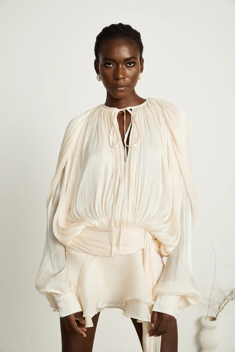 Close-up of a woman in a cream long sleeve tie neck mini dress, showcasing the pleated bodice and soft fabric texture, with pearl earrings.