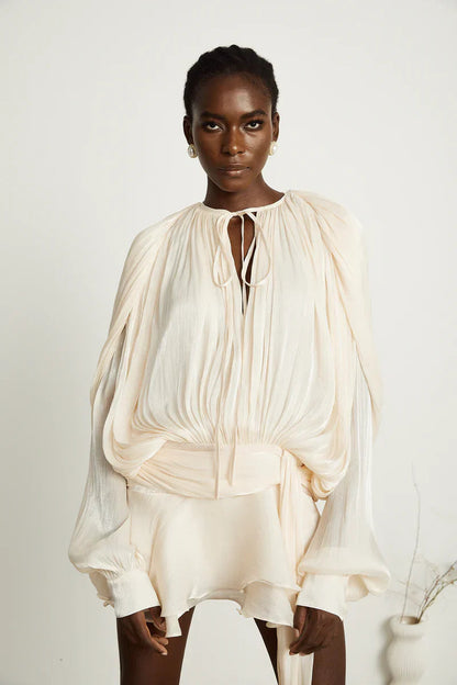 Close-up of a woman in a cream long sleeve tie neck mini dress, showcasing the pleated bodice and soft fabric texture, with pearl earrings.