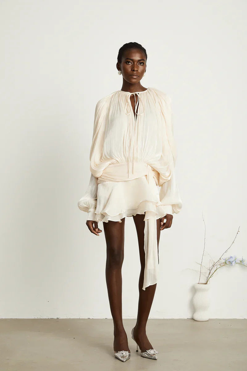 Full body shot of a woman in a cream long sleeve tie neck mini dress with a cinched waist belt and ruffled hem, posing with white embellished heels.