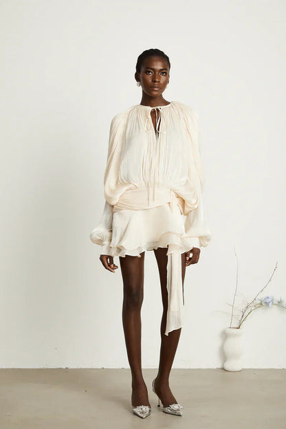 Full body shot of a woman in a cream long sleeve tie neck mini dress with a cinched waist belt and ruffled hem, posing with white embellished heels.