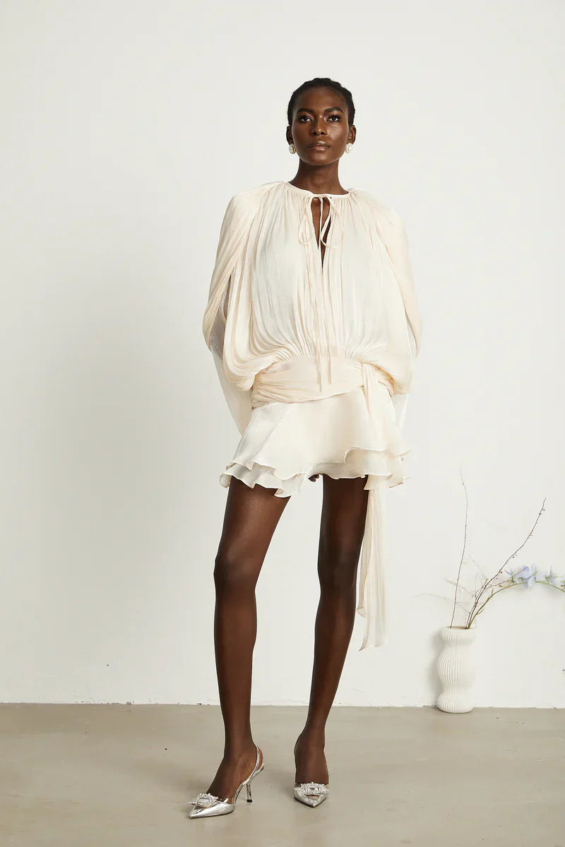 Woman standing in a cream long sleeve tie neck mini dress, featuring a flowing skirt and a long belt tie, paired with sparkling white slingback heels.