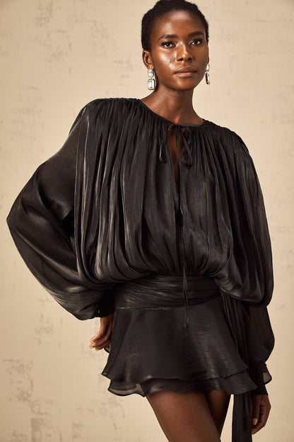 Close-up of a woman wearing a black long sleeve tie neck mini dress, highlighting the pleated fabric and oversized sleeves, with statement silver earrings.