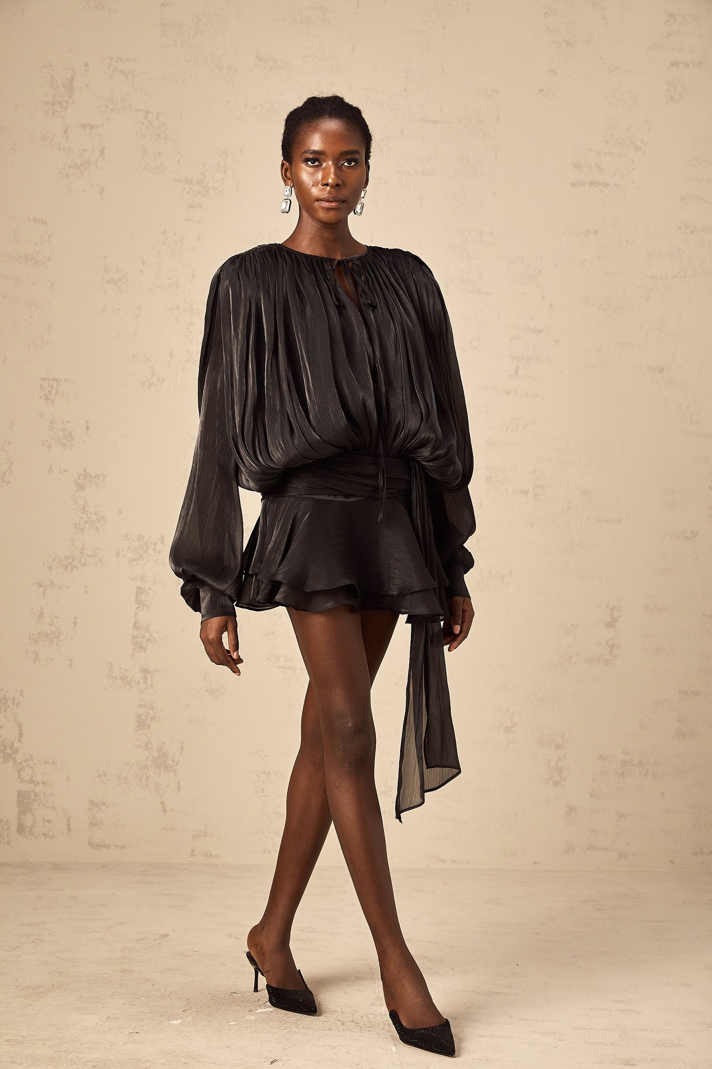 Woman in a black long sleeve tie neck mini dress with a flowing silhouette, posing elegantly against a light textured wall with black pointed heels.