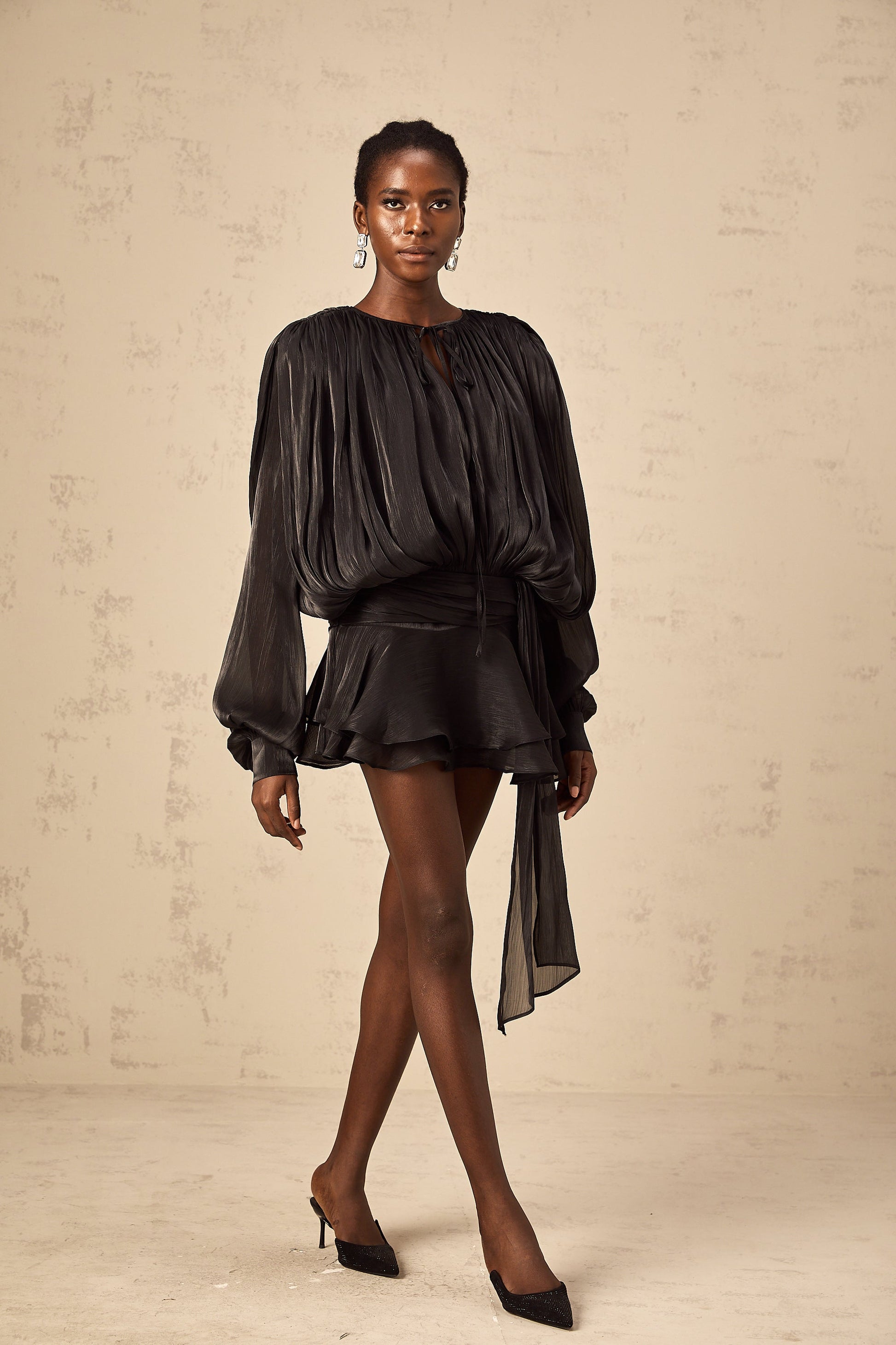 Woman in a black long sleeve tie neck mini dress with a flowing silhouette, posing elegantly against a light textured wall with black pointed heels.