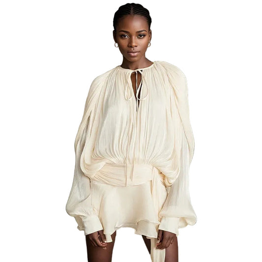 Front view of a woman wearing a cream long sleeve tie neck mini dress with a wide belt and voluminous sleeves, against a dark textured background.