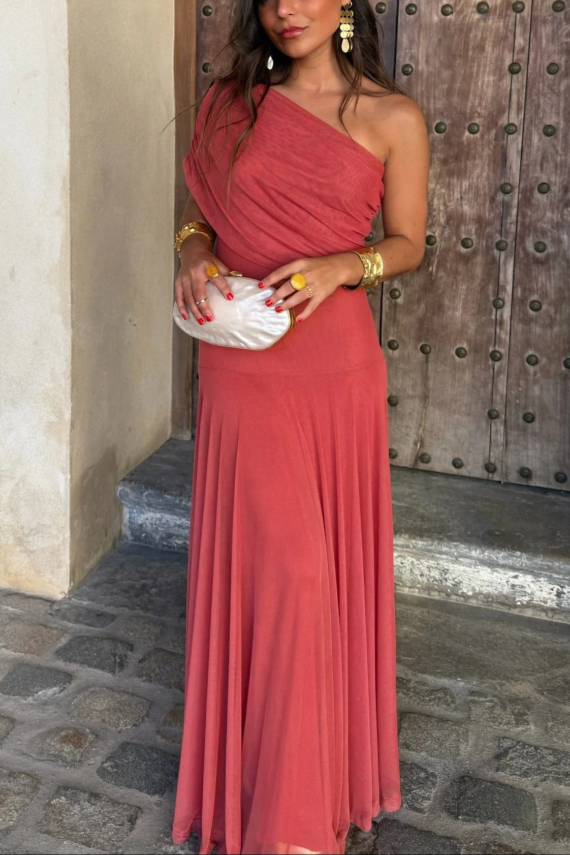 Full view of a women's coral pink off-shoulder draped mesh maxi dress, featuring a one-shoulder style and a long, flowing skirt.