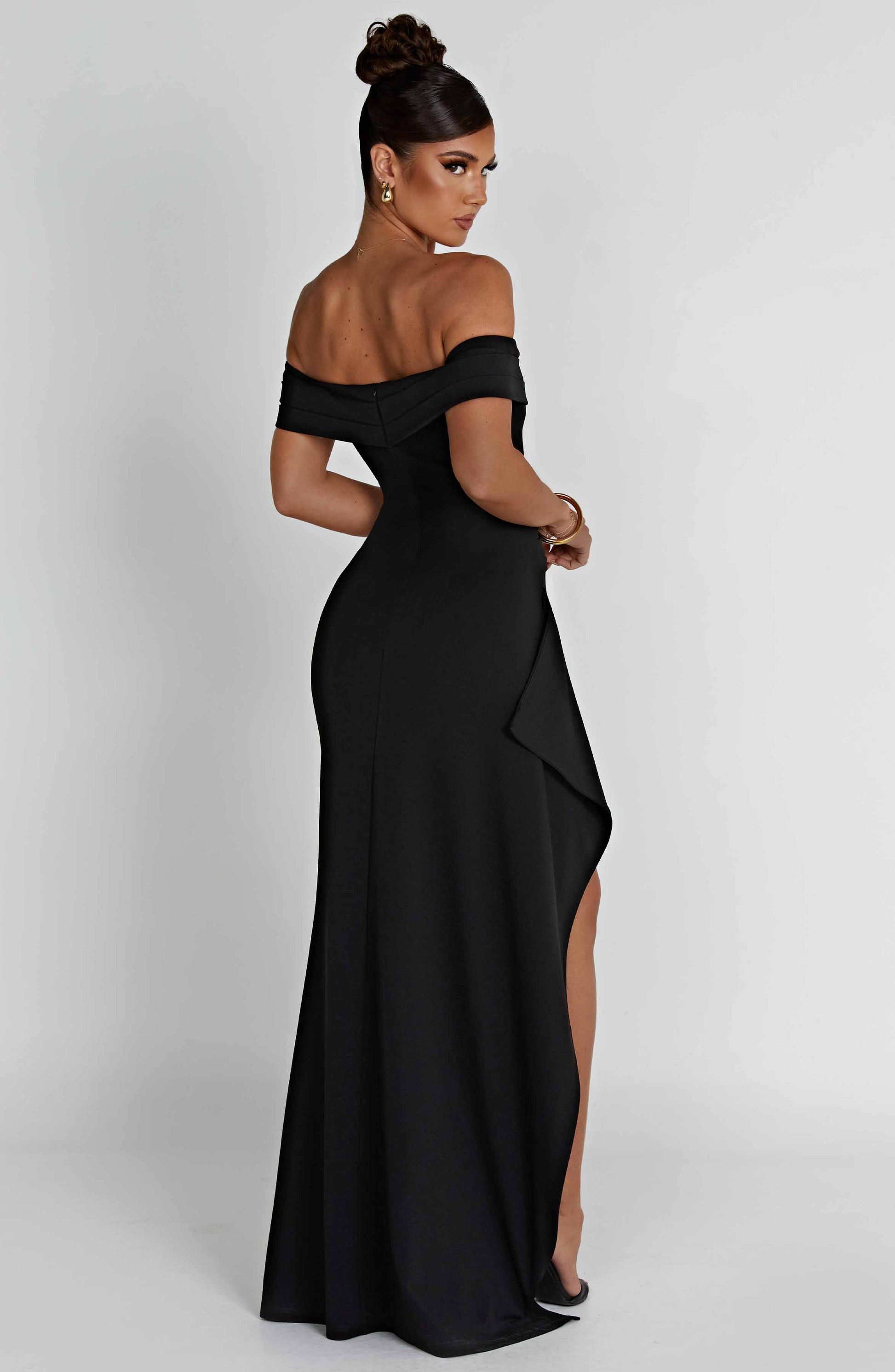 Back three-quarter view of the black off-shoulder pleated maxi dress, highlighting the high slit and draped accent.
